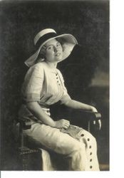 Unidentified young woman seated in chair with large summer hat and linen-like long dress with buttons down front, about 1913