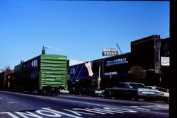 Train down main traveling north on North Main, passing Medico Drugs and Tombe Reality, 1977