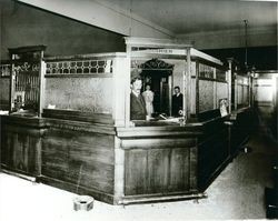 Three bank employees are standing behind a cashier's window inside the Analy Savings Bank, 1904