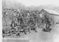 Woods crew or sawmill crew standing and seated in front of rail tracks and cabins or houses, Jenner, California in background