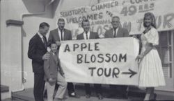 Six people standing in front of the Chamber of Commerce building holding sign celebrating the 15th Annual Apple Blossom Tour, about 1962