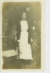 Unidentified young woman in dress, coat, hat and gloves in garden scene with flowers and lattice fence