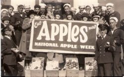 Group of naval officers and sailors and women around a sign "Eat Apples now National Apple Week"