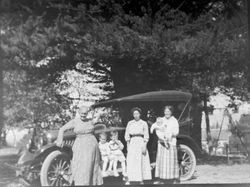Group of three women and three children pose in front of a vintage car, about 1915