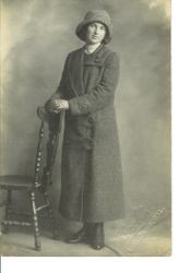 Unidentified young woman in long coat and hat standing by chair