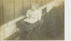 Girl baby in white dress seated in wagon in grass next to a house, about 1912