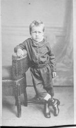Unidentified young boy, about 4 years old standing with legs crossed next to a Victorian chair in a studio photo, about 1860s