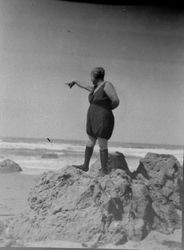 Woman in a 1920s bathing suit stands on some rock by the sea shore and is pointing out to sea