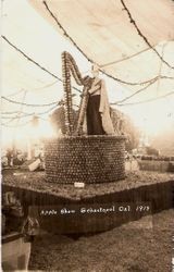 Gravenstein Apple Show display of Round apple covered structure with harp made of apples being played by King Gravenstein mannequin dressed in cape made of apple slices