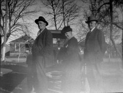 Three unidentified older adults, one woman between two men stand in front of a car, possibly, about 1920s