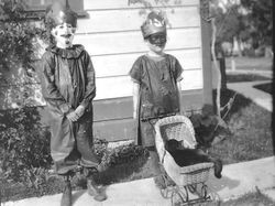 Two children, a boy and a girl, in costumes and masks and stand beside a house on the sidewalk, 1920s