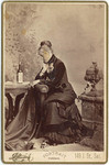 [Seated woman, wine bottle and glass on small table at left]