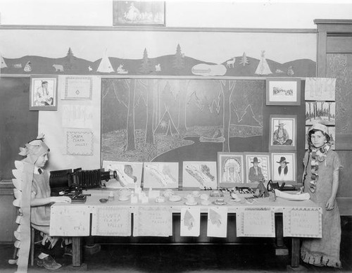 Two children in Native American dress beside classroom displays