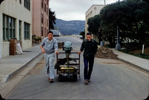 Divers James B. Jordan and Earl D. Squier pulling a cart with scuba diving tanks and gear, and a boat engine, on the campus of the Scripps Institution of Oceanography