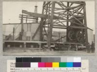 The base of a 122' oil well derrick showing bull wheels to the right and also the walking beam supported on Sampson post and operated by Ironwood Pitman. The engine is enclosed in the little house. Picture was taken during pumping operations. Bakersfield, California. Dec. 1927