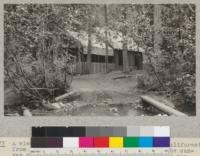 A view of the kitchen and mess hall at Camp Califorest from across Schneider Creek, looking toward the canvas covered mess portion in the foreground and the kitchen under the shingle roof in the background