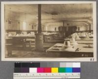 The interior arrangement of the cookhouse. The men are seated at small tables, eight at a table, and are arranged as much as possible in crews so that when dinner has to be sent out to the woods a whole table is missing, rather than one here, one there. The kitchen is at the end