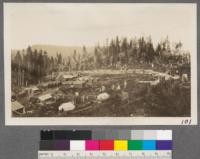 GR 118. Grays River, Washington camp of the Portland Lumber Company. To the right the family houses are arranged in a long line. Those unable to get houses live in the tents in the foreground. In this camp there are 25 families. The policy of this company is to get married men whenever possible. They are more steady. In the left portion of the photo the largest building is the cookhouse. Just beyond it in order lie the commisary, the company house where visitors stay and the office men live, a family house, the superintendent's cottage and the engineering shack. On this side of it are the bunk houses. Those on the same side of the track are for 16 men each. On the opposite side are four for eight men each and 8 small shacks for two men each. Across from the cookhouse is the barber shop and bath house, only the top of which can be seen. In the foreground on the same side of the track as the cookhouse is the filing room and the sane house, on the opposite side, the tool house. In the extreme left just the corner of the shops and engine sheds show. The carpenter shop is large enough to make new cars if need be and this winter they practically rebuilt one of the locomotives in the machine shop. The camp on the whole is better than the average and in most respects is thoroughly modern