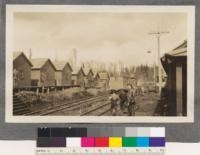 The thoroughfare of the Rinearson camp of the Portland Lumber Company. On the left is the row of bunk houses, each holding 8 men. On the right in the background can be seen the porch of the commissary. The photo was taken just after dinner when the men were going over to the "mulligan" car to go back to the woods. This car, which with the locomotive, is standing in front of the commissary is an old box car with four benches running lengthwise. In level country it is a good scheme, particularly when rainy, but the men have to crowd in so tightly that where rough it is dangerous and in the other camps of the company, the men are carried on flat cars where they have a chance if anything happens