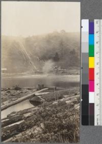 Showing method of getting logs from cars into pond, where the two are at much different elevations. Logs are unloaded at top from cars by means of hook and cable operated by small spool donkey. Logs slide down by gravity into pond below and are poled to the mill. Caspar Lumber Company, Caspar, California. May, 1920. E.F