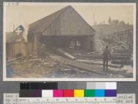 Log slip - Clio mill, Two log joined by coupler pulled up with cable and bull wheel - the cable is pulled out by the pond man and log deck man. Note man on burner conveyor. Boiler house at left