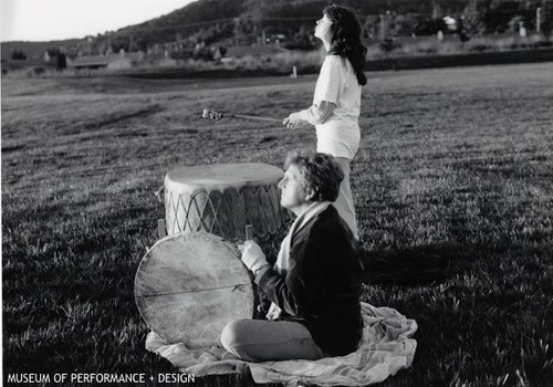 Two individuals outdoors with drums, one sitting, one standing (related to a performance of "Circle the Earth")