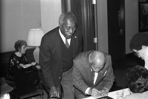 Nate Holden watching Gil Lindsay sign a paper at an event, Los Angeles, ca. 1984