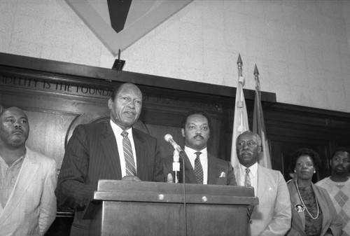 Jessie Jackson and Nate Holden standing with Tom Bradley speaking at City Hall, Los Angeles, 1988