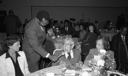 Dr. H. Claude Hudson sitting at a table as he speaks into a microphone, Los Angeles, 1983