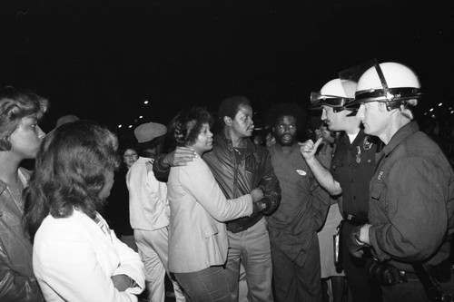 White police officers in riot gear talking with a distressed African American couple among a crowd, Los Angeles, 1980