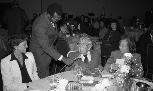 Dr. H. Claude Hudson sitting at a table as he speaks into a microphone, Los Angeles, 1983