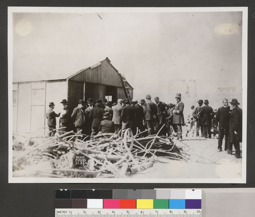 [Crowd gathered outside building. Fire-damaged scrap metal, foreground.]