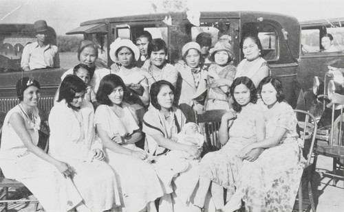 Christening party for Adeline Cabreana, Guadalupe Produce Labor Camp, Lompoc : 1932. Seated L-R: Margarita Bautista, unidentified, Maria Salutan, Mati holding the baptized Adeline, Maria Tenequez Cabreana, and Margarita Cabatuan. Standing: Maria Putin, Insay Mansalvas, Justa Abenida, unidentified, Maria Completo, and Bernalda Tenequez. In the driver's seat: Sarapio Cabatuan, nearest car; Santiago Salutan, distant car