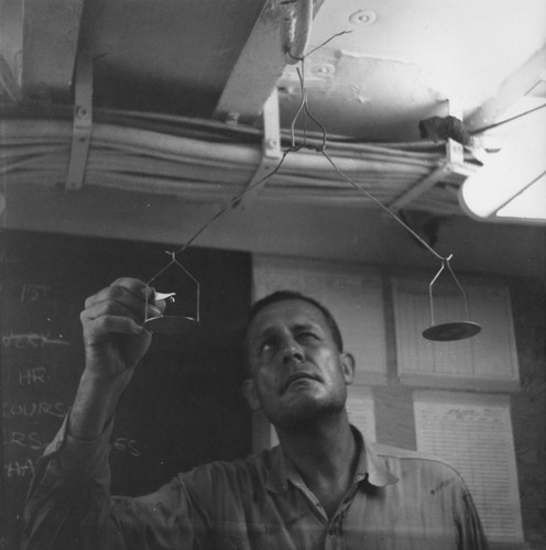 Roger Revelle and his makeshift scale, shown here adjusting the balance arm for 1 gram, aboard R/V Spencer F. Baird (ship) while on the Capricorn Expedition (1952-1953). November 1952