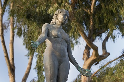 Standing: detail view of bronze figure with hands outstretched and eucalyptus trees in the background