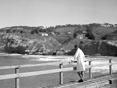 A unidentified person standing on the Scripps pier enjoying a view of the ocean and the coastline. Note the cottages and trestle walk bridge in the background at Scripps Institution of Oceanography. Circa 1950