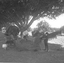 View of a family in a garden. Chickens strut around. A man is reclined in a barber chair, a boy plays with a train set