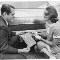 Caption reads: Lt. Governor Ed Reinecke helps Mrs. Jessica Bradley attach a new gold on blue license plate to her car."