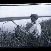 A young men sitting in cattails holding a shotgun