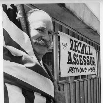 Jack Hickman, husband of Dr. Irene Hickman, an osteopath and the Sacramento County assessor, draped in American flag next to "Recall Assessor" sign