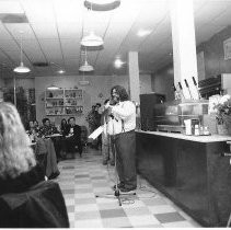 Gene Black gives presentation at microphone at Java City restaurant during WordJam event. Contestants lined up behind him. Audience seated at tables