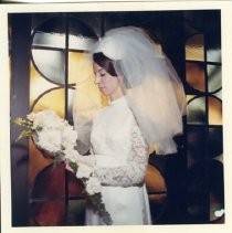 Bride Candace Lusk Smallwood standing in silhouette in front of stained glass windows. She is looking at a white flower bouquet