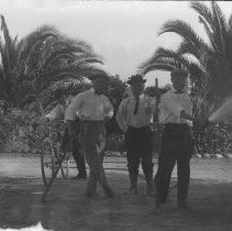 Exterior view of four men in front of a palm tree one with a garden hose, one with a rifle and the one behind with a bicycle
