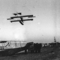 Two bi-planes; one on a beach and the other flying overhead