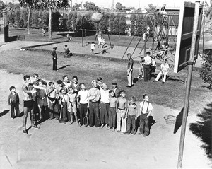 A group of young men line up to shoot some basketballs, while other kids play on the swing set in the background