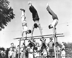 Three men practicing gymnastics on the parallel bars, as many young onlookers cheer them on