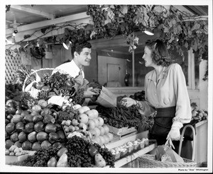 A woman wearing a palm tree brooch is being assisted by a man working at the produce stand at the Farmers Market