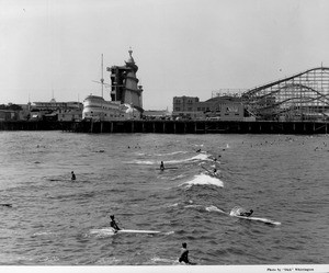 People ride surf boards and paddle boards in front of an amusement park