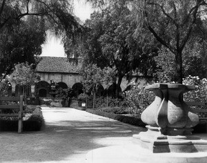 Two women in historical costumes stand in front of a fountain in the San Fernando Mission