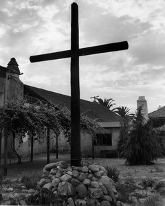 A large wooden cross standing in the garden of the San Gabriel Mission, with grape vines and one of the buildings in the background